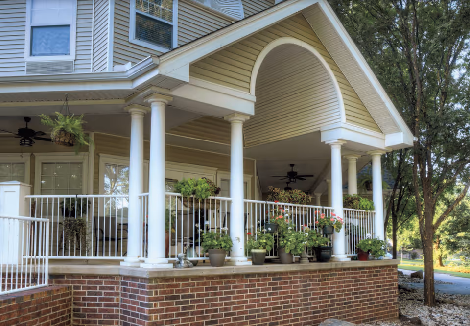 Front porch of a senior living building with white columns, railing, and potted plants.