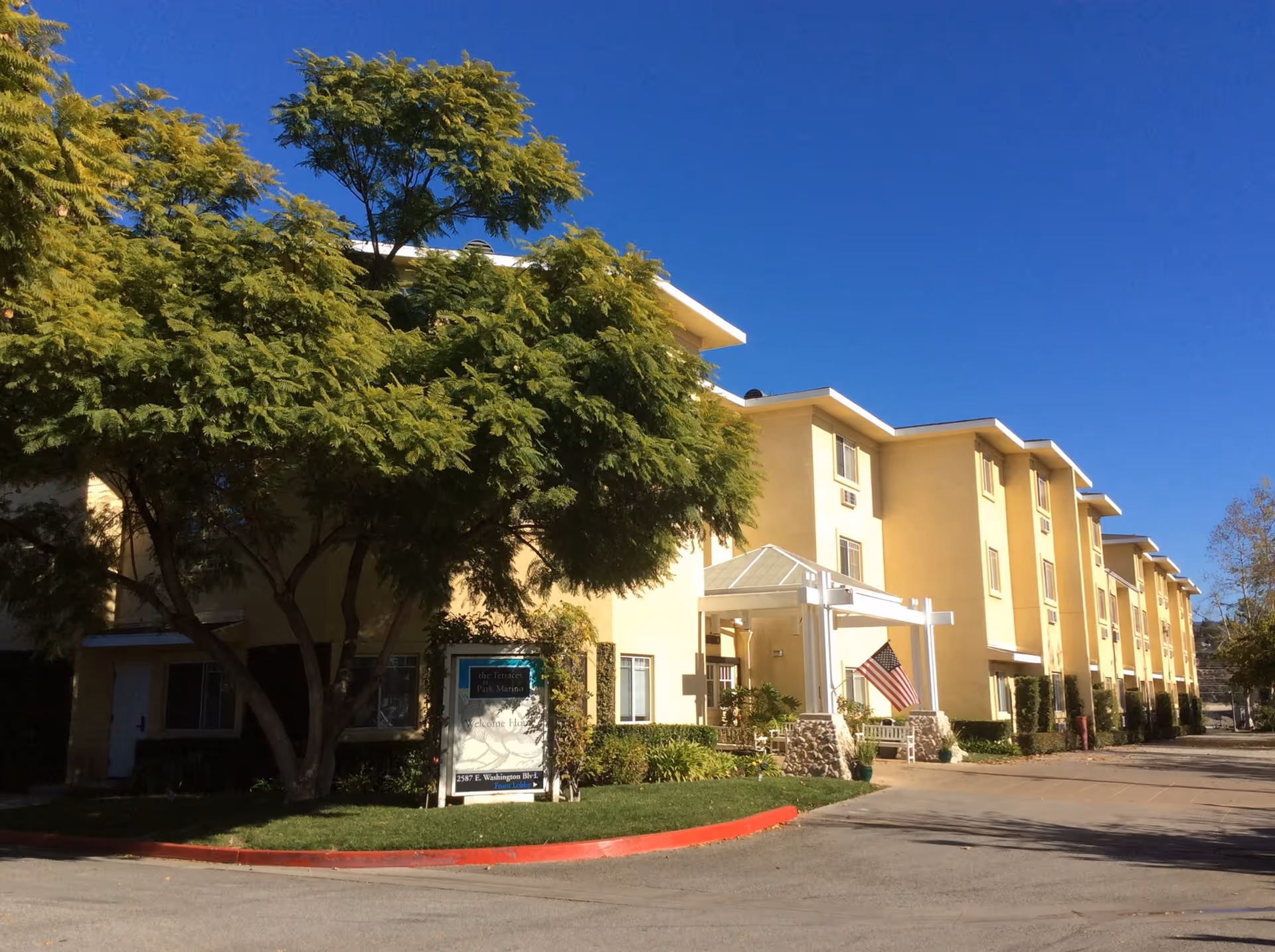 Yellow multi-story senior living building with an entrance canopy, American flag, trees, and a clear blue sky.