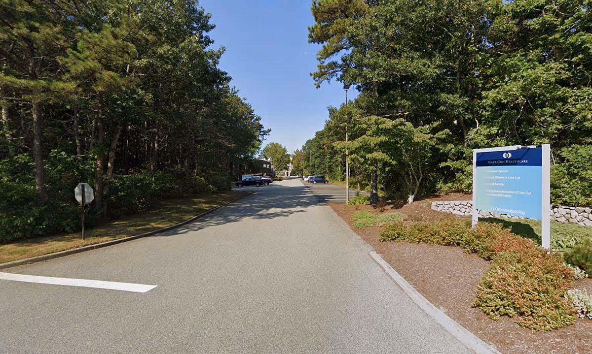 A tree-lined driveway leads to a facility entrance with a blue sign on the right.
