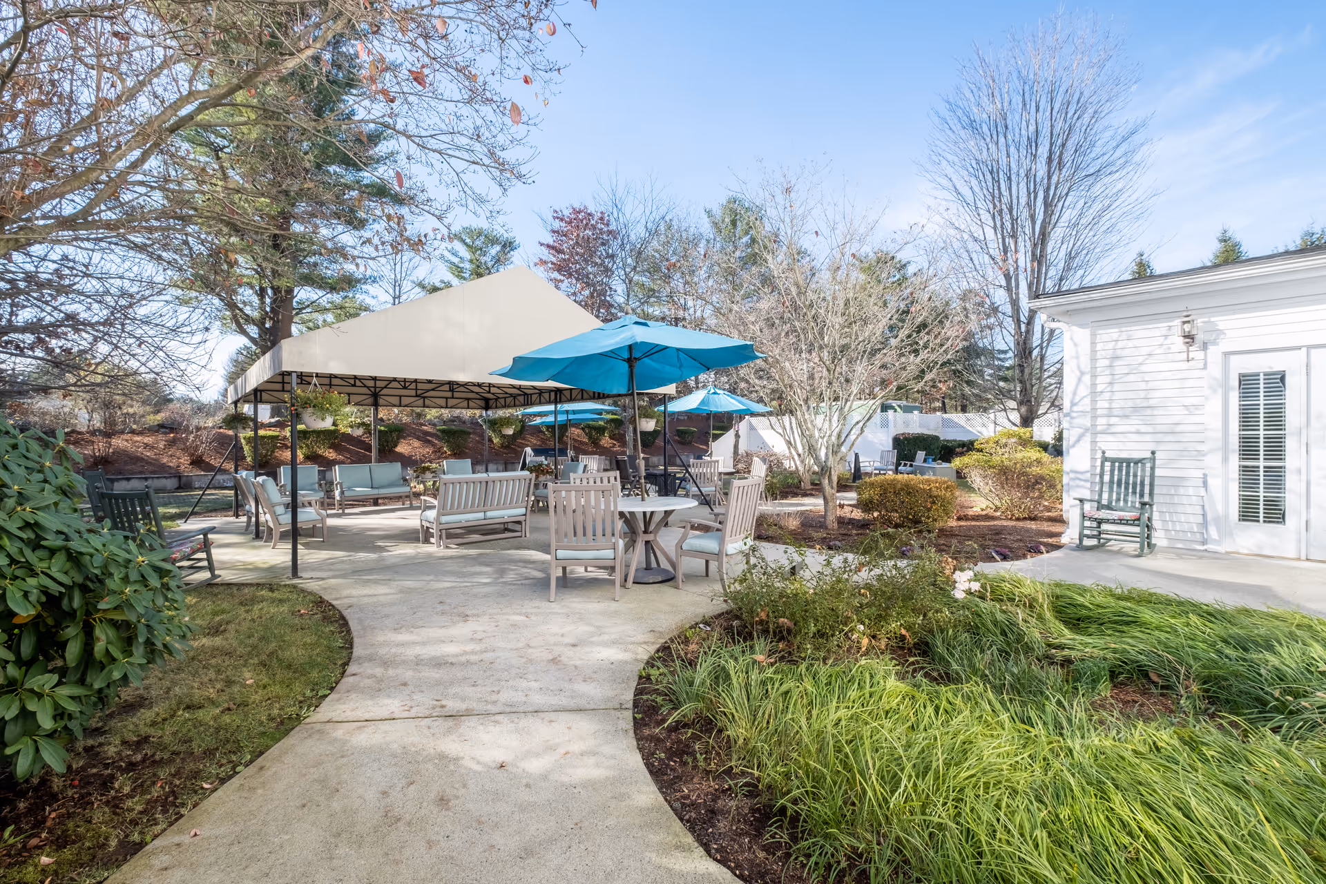 Outdoor courtyard with tables, chairs, umbrellas, and a canopy near a white facility building.