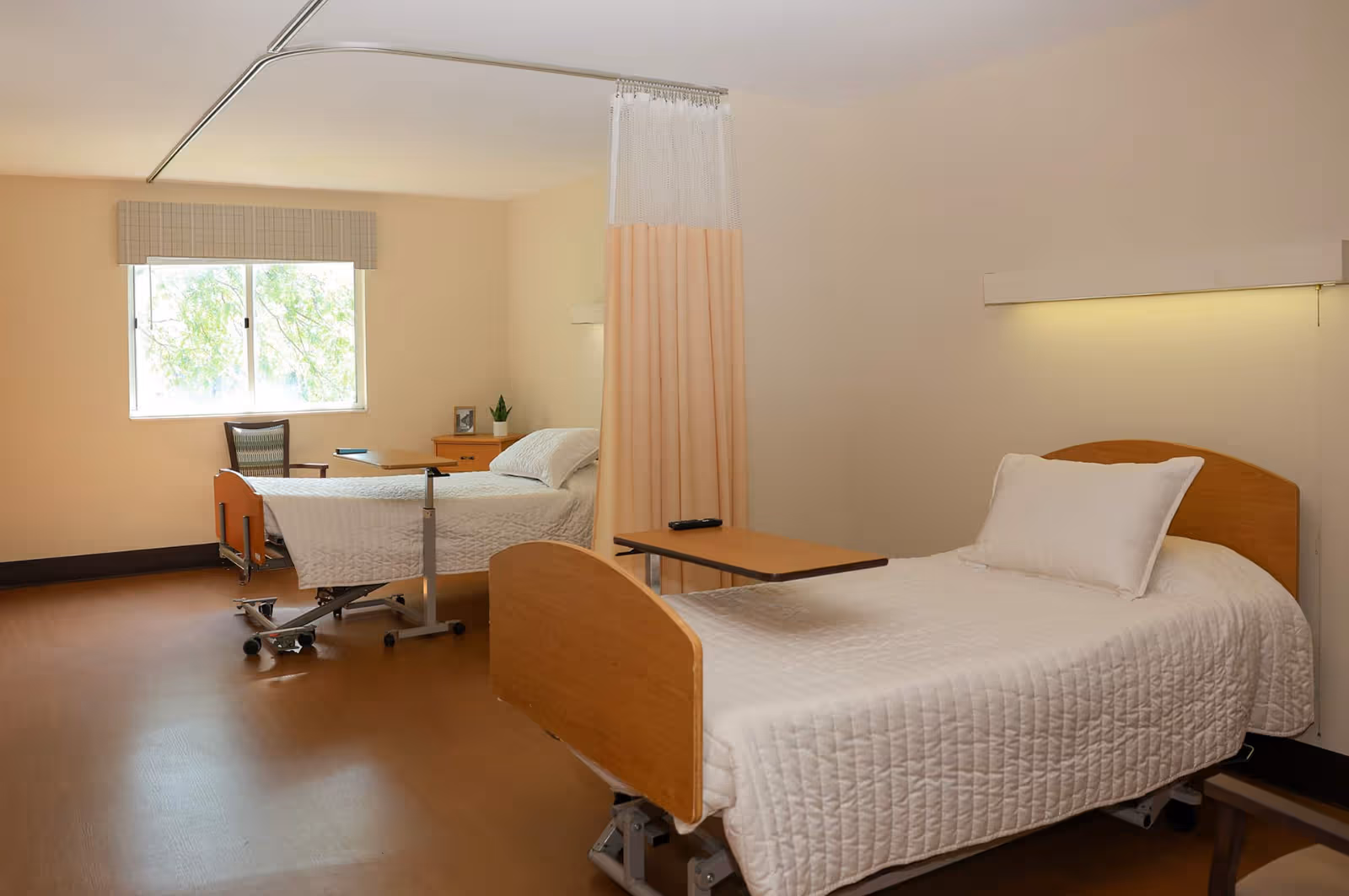 Two hospital beds with a privacy curtain, a window, and simple furniture in a bright room.
