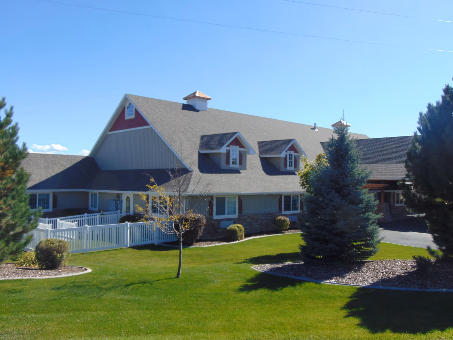 Large gabled memory care building with a white picket fence, green lawn, and trees in front.