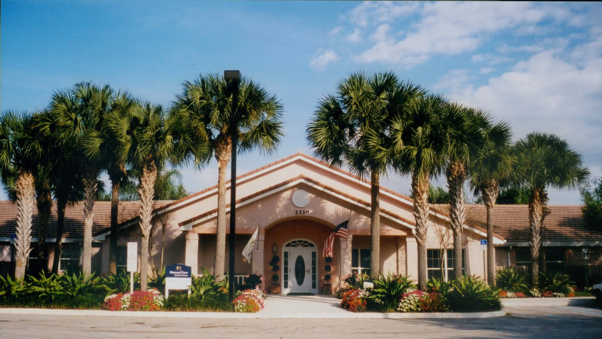 Front exterior view of Arden Courts - ProMedica Memory Care Community in West Palm Beach, featuring a single-story building with a peaked roof entrance, surrounded by palm trees and landscaped greenery under a partly cloudy sky.