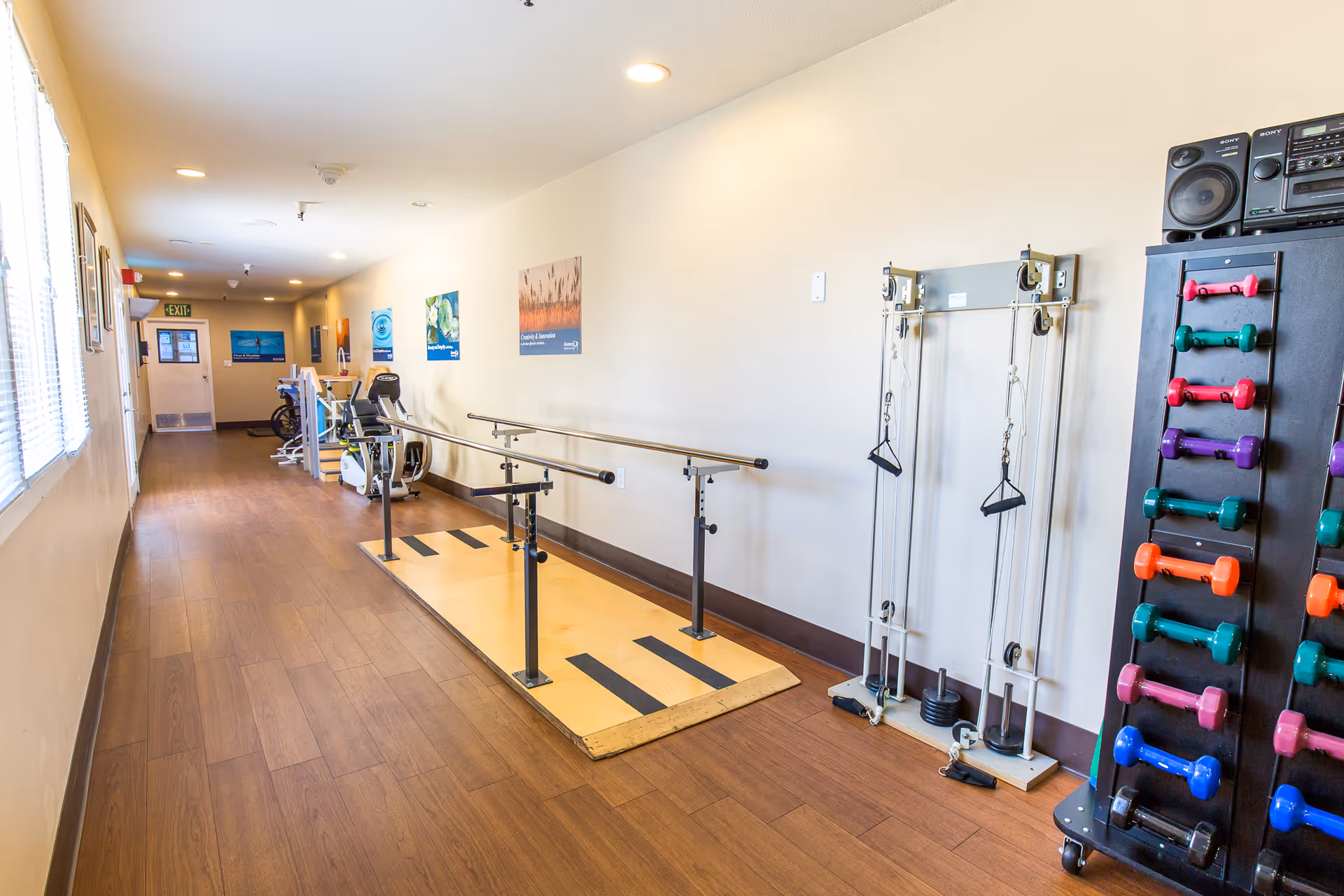 A hallway in a senior living facility equipped with exercise equipment including parallel bars for walking practice, a weight machine, and a rack of colorful dumbbells. The floor is wooden, and the walls are decorated with motivational posters. There is a window on the left side letting in natural light.