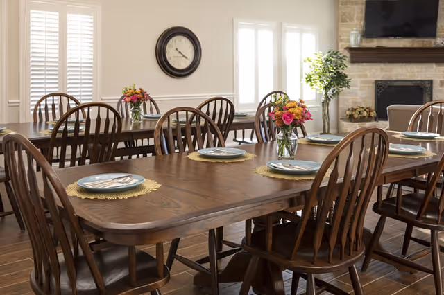 A dining room with two wooden tables set with plates, silverware, and floral centerpieces. The room features wooden chairs, a wall clock, large windows with white shutters, a stone fireplace, and a potted plant near the fireplace.