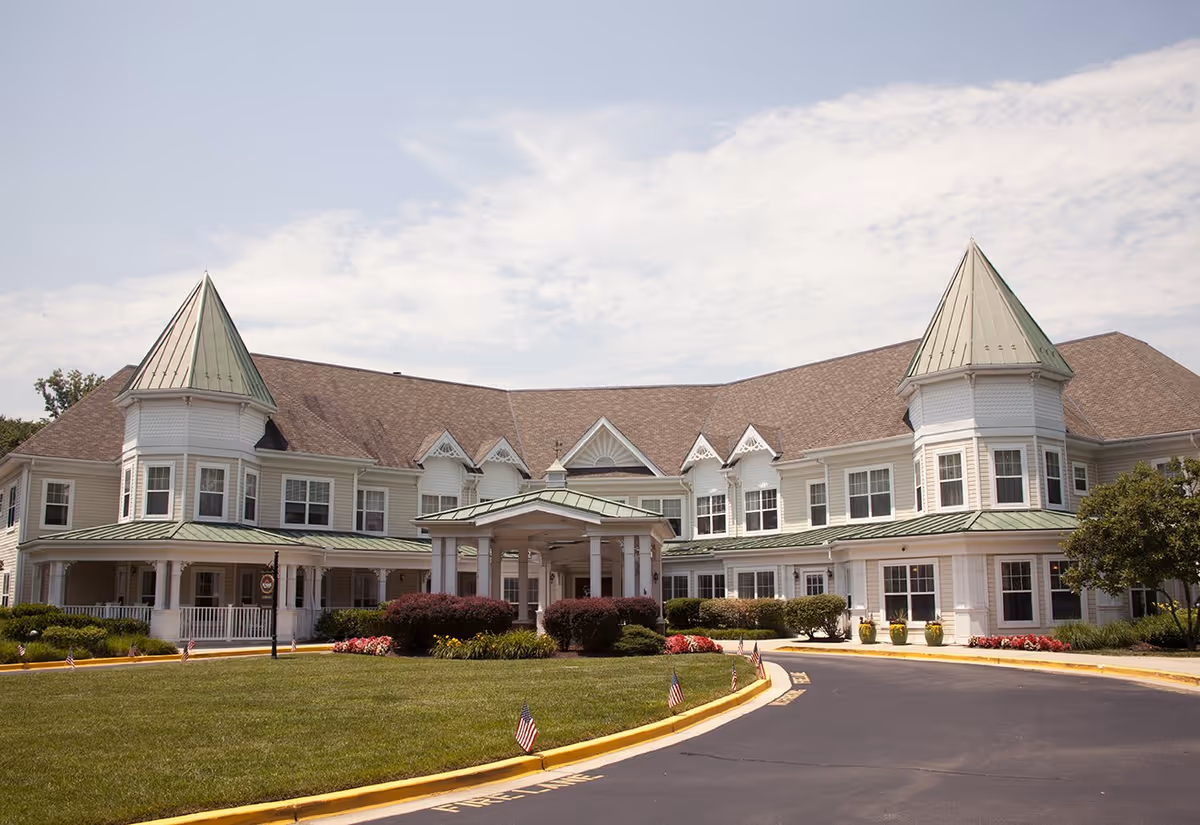Front entrance of a large two-story white senior living building with green-roofed turrets, a covered portico, and a landscaped lawn and driveway.