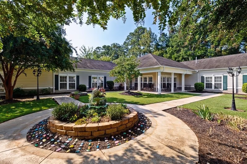 Outdoor courtyard area at Oaks at Greenwood featuring a circular raised garden bed with various plants and colorful painted stones around its base, surrounded by paved walkways, green grass, trees, and a single-story building with a covered porch and rocking chairs.