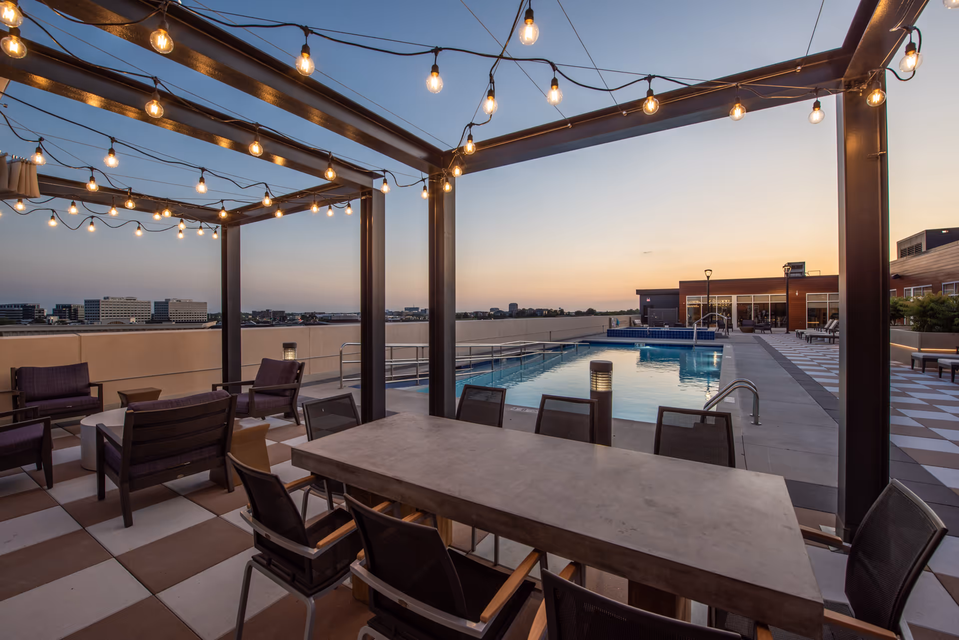 Outdoor rooftop patio area at sunset with string lights hanging from a pergola, a concrete dining table with chairs, lounge seating, and a swimming pool with a ramp and handrails. Buildings and cityscape are visible in the background.
