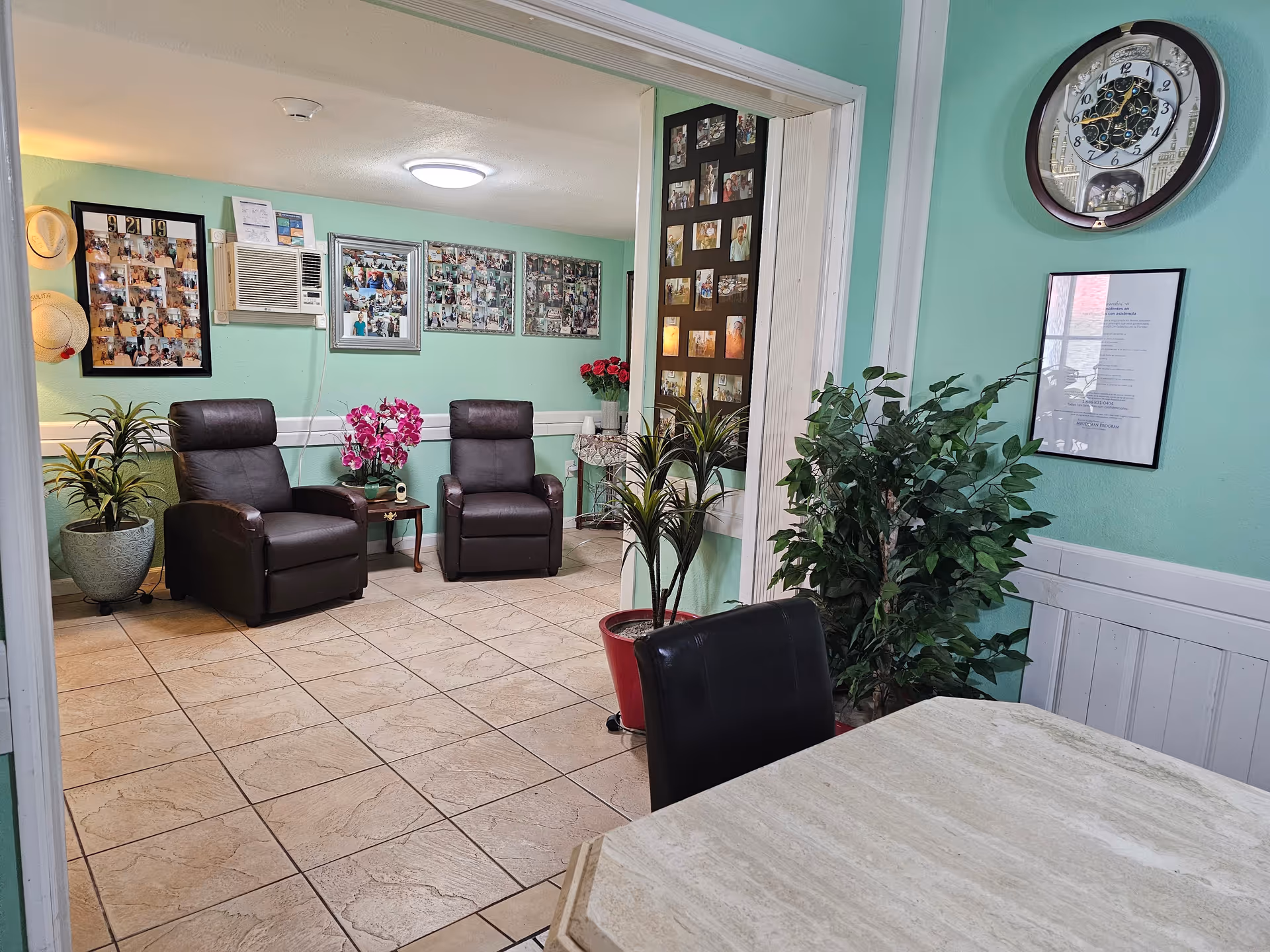 A pastel-green senior living lounge with leather chairs, plants, and photo collages on the walls.