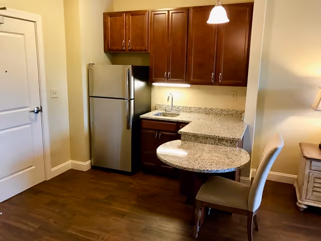 Small kitchen area with wooden cabinets, a stainless steel refrigerator, a granite countertop with a built-in sink, and a round granite breakfast bar with a beige upholstered chair. The floor is wooden, and there is a white door to the left and a small wooden side table with a lamp to the right.