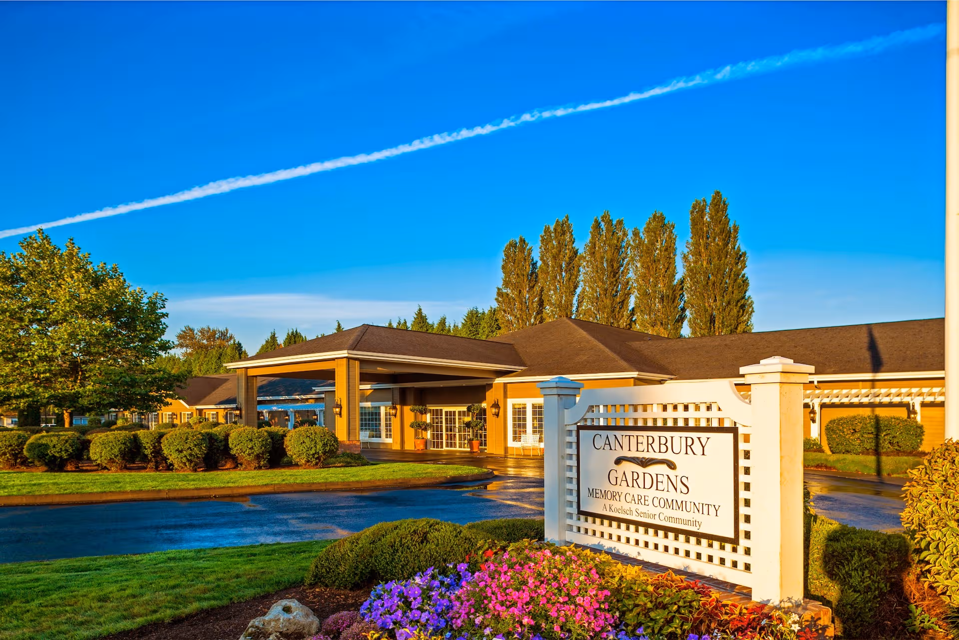 Exterior view of Canterbury Gardens Memory Care Community entrance with landscaped sign and bright blue sky.