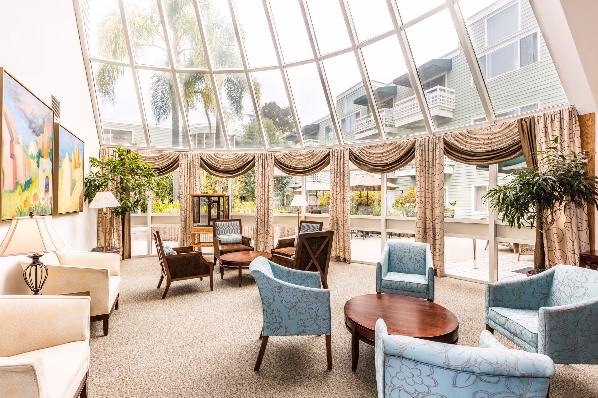 Bright and spacious lounge area with large curved glass windows and ceiling, allowing natural light to fill the room. The room is furnished with multiple armchairs in blue and beige tones arranged around wooden coffee tables. There are two large paintings on the left wall, potted plants near the windows, and beige patterned curtains framing the windows. Outside, a patio area with tables and umbrellas is visible.