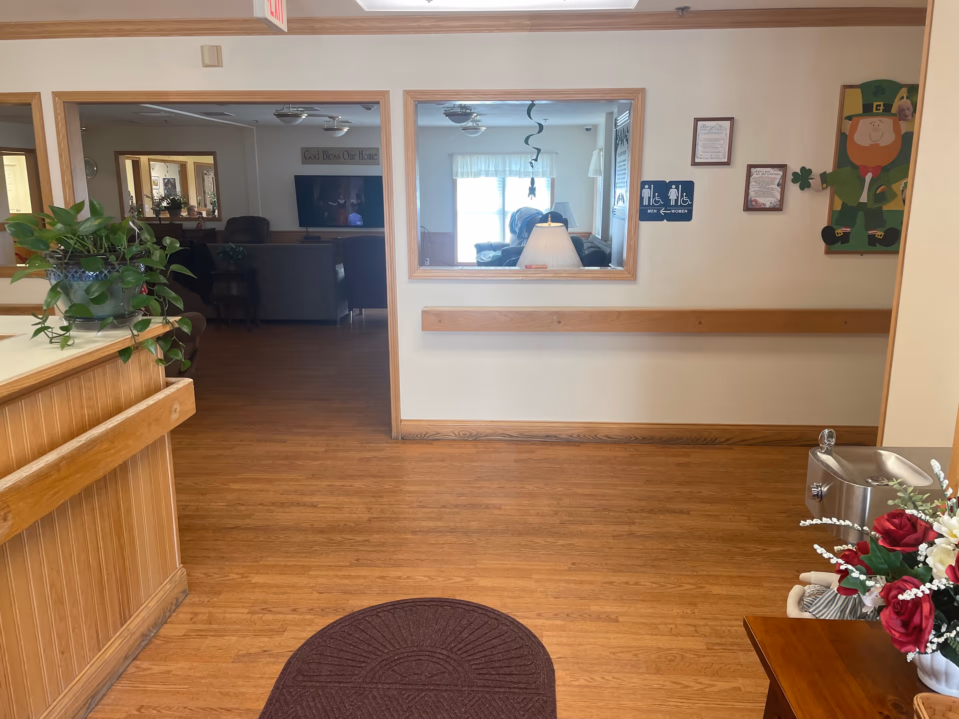 Interior hallway of Serenity Assisted Living with wooden floors and light-colored walls. There is a wooden reception counter with a green plant on it to the left, a water fountain and a table with a flower arrangement to the right. A window in the wall shows a room with a lamp and seating area. Signs indicating restrooms for men, women, and accessible facilities are visible on the wall.