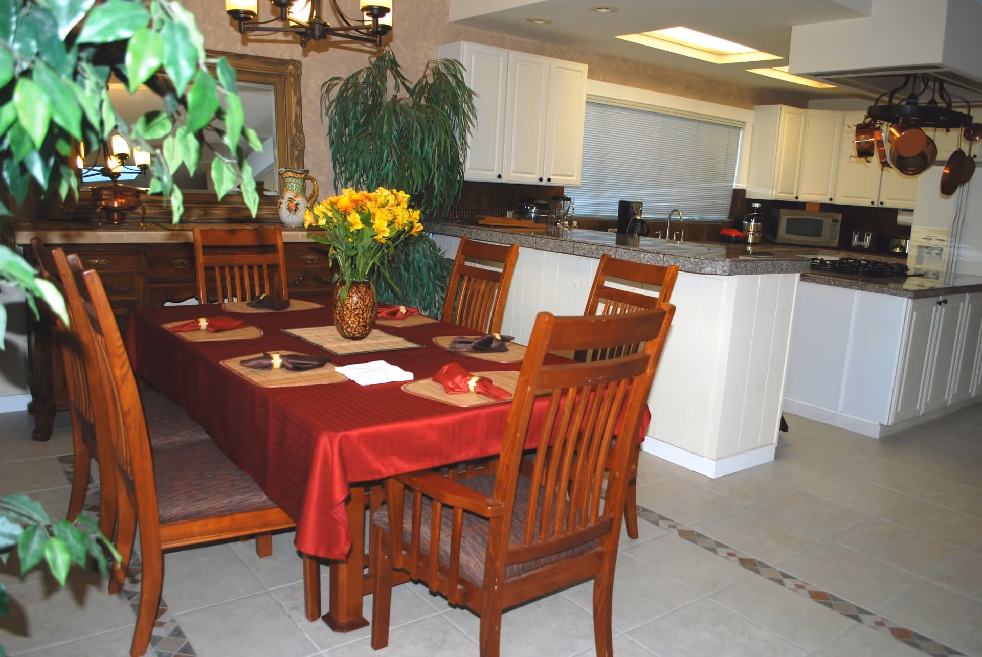 A dining room scene with a red tablecloth, wooden chairs, and a kitchen backdrop.