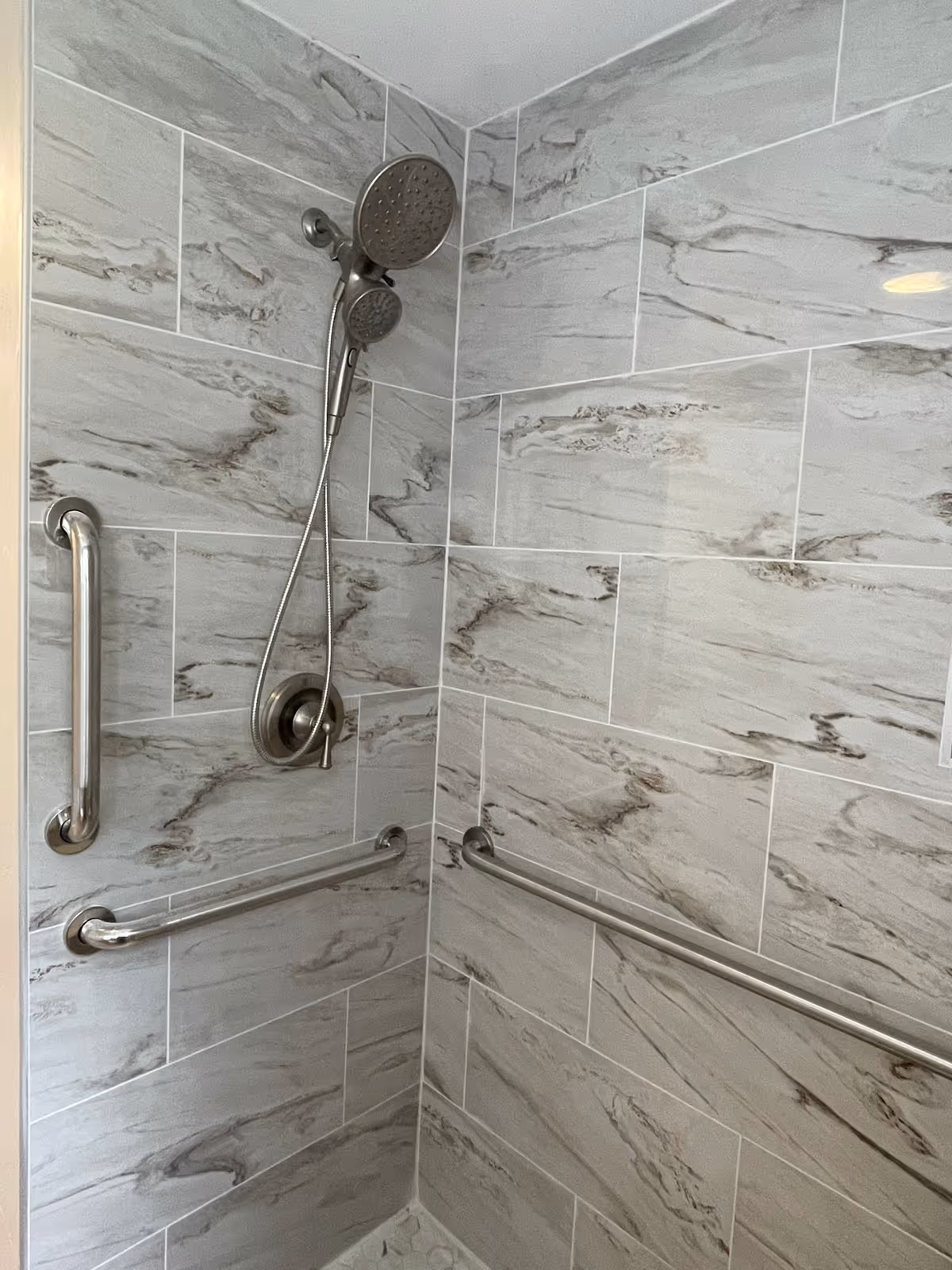 Shower with marble-pattern tiles, a handheld head, and multiple grab bars.