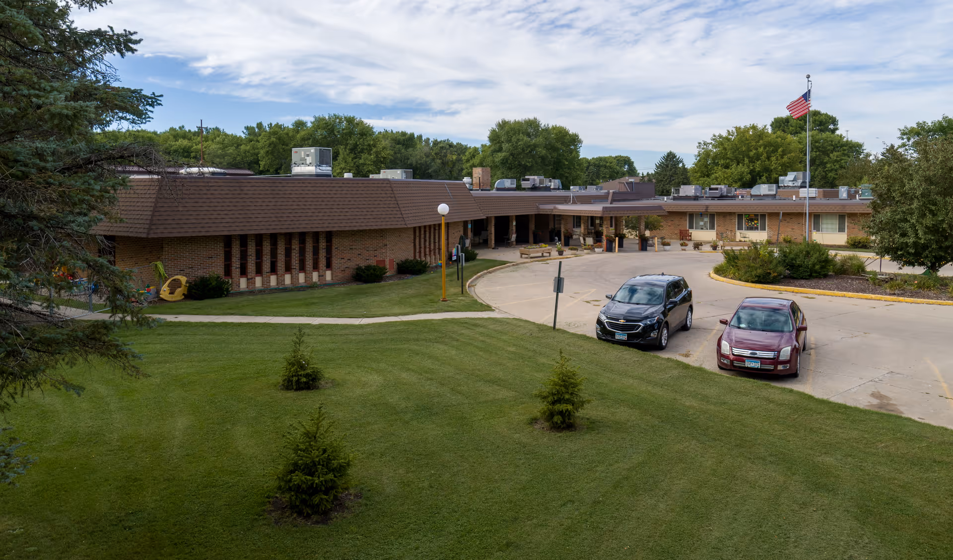 Exterior view of Morris Health Services DBA West Wind Village, showing a single-story brick building with a brown roof, a curved driveway with two parked cars, a flagpole with an American flag, and a well-maintained lawn with small trees and shrubs under a partly cloudy sky.