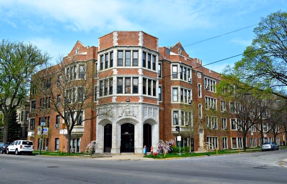 Exterior view of a large, multi-story brick building with many windows and an arched entrance. The building is situated on a street corner with trees and parked cars nearby under a blue sky with some clouds.