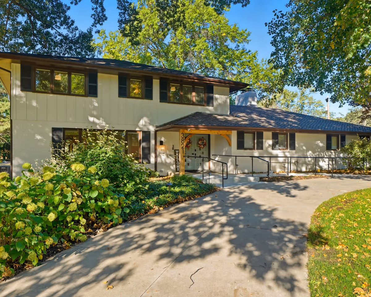 Two-story senior housing building entrance with ramp, greenery, and sunny walkway.