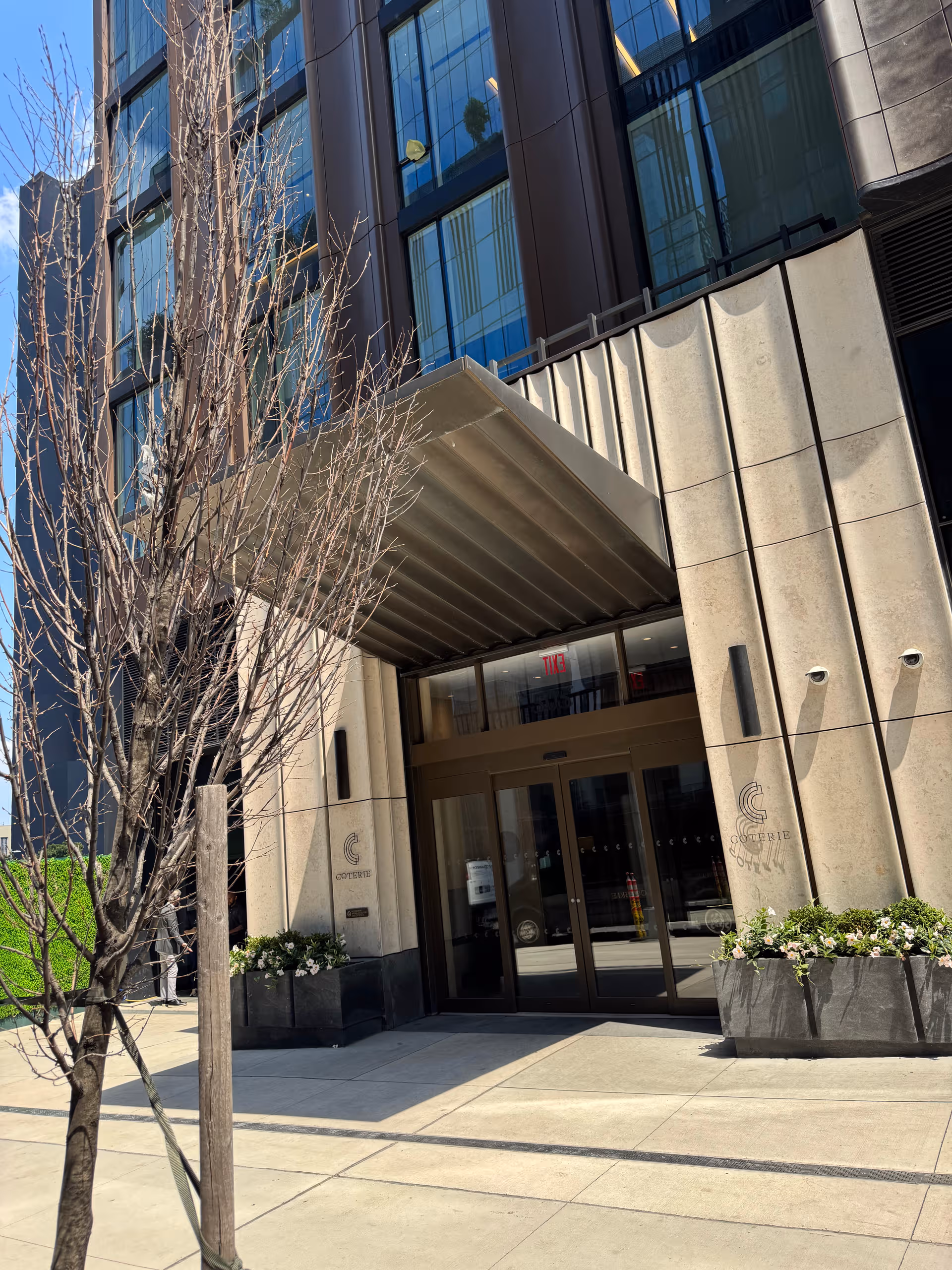Modern building entrance with glass doors, concrete columns, and planters at the Coterie Hudson Yards.