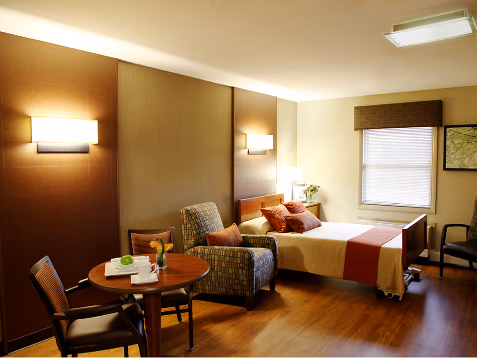 A cozy bedroom in a senior living facility with a single bed covered in beige and orange bedding, a patterned armchair, a small round wooden table with two chairs, and a window with blinds. The room has warm lighting from wall sconces and a table lamp, wooden flooring, and a framed picture on the wall.