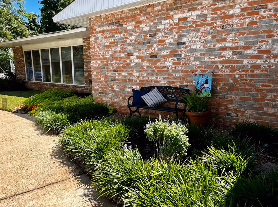 Brick exterior of a senior living facility with a bench, potted plant, decorative sign, and landscaped greenery along a sidewalk.