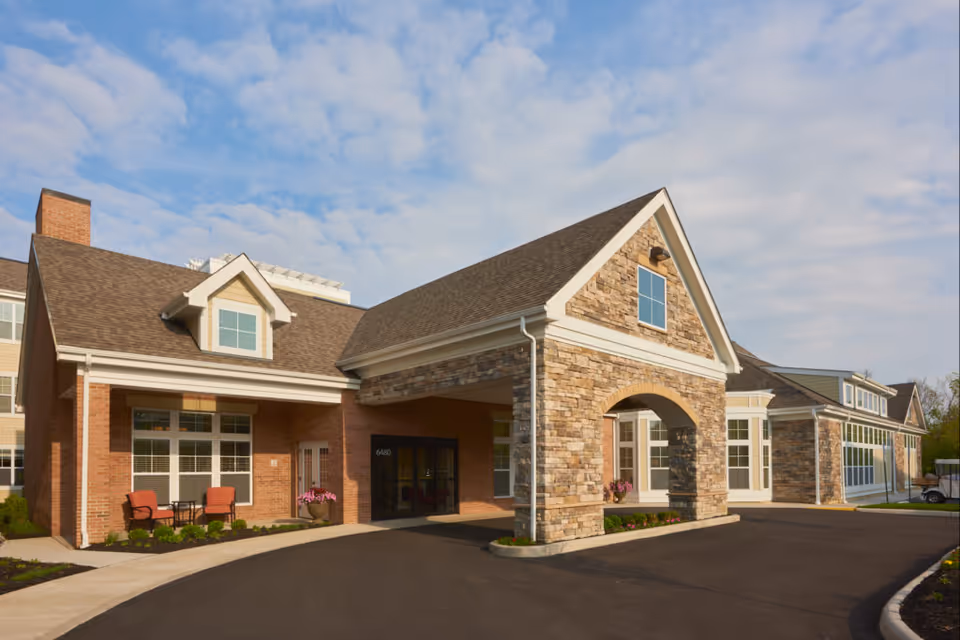 Front entrance of a brick and stone assisted living facility with a covered porte-cochere and outdoor seating by the windows.