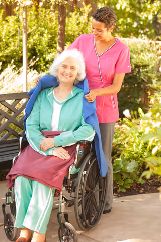 Caregiver in pink scrubs helps an elderly woman in a wheelchair outdoors in a garden.