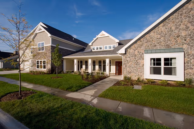 Stone-and-siding senior living building with a covered porch, walkway and manicured lawn under a blue sky.
