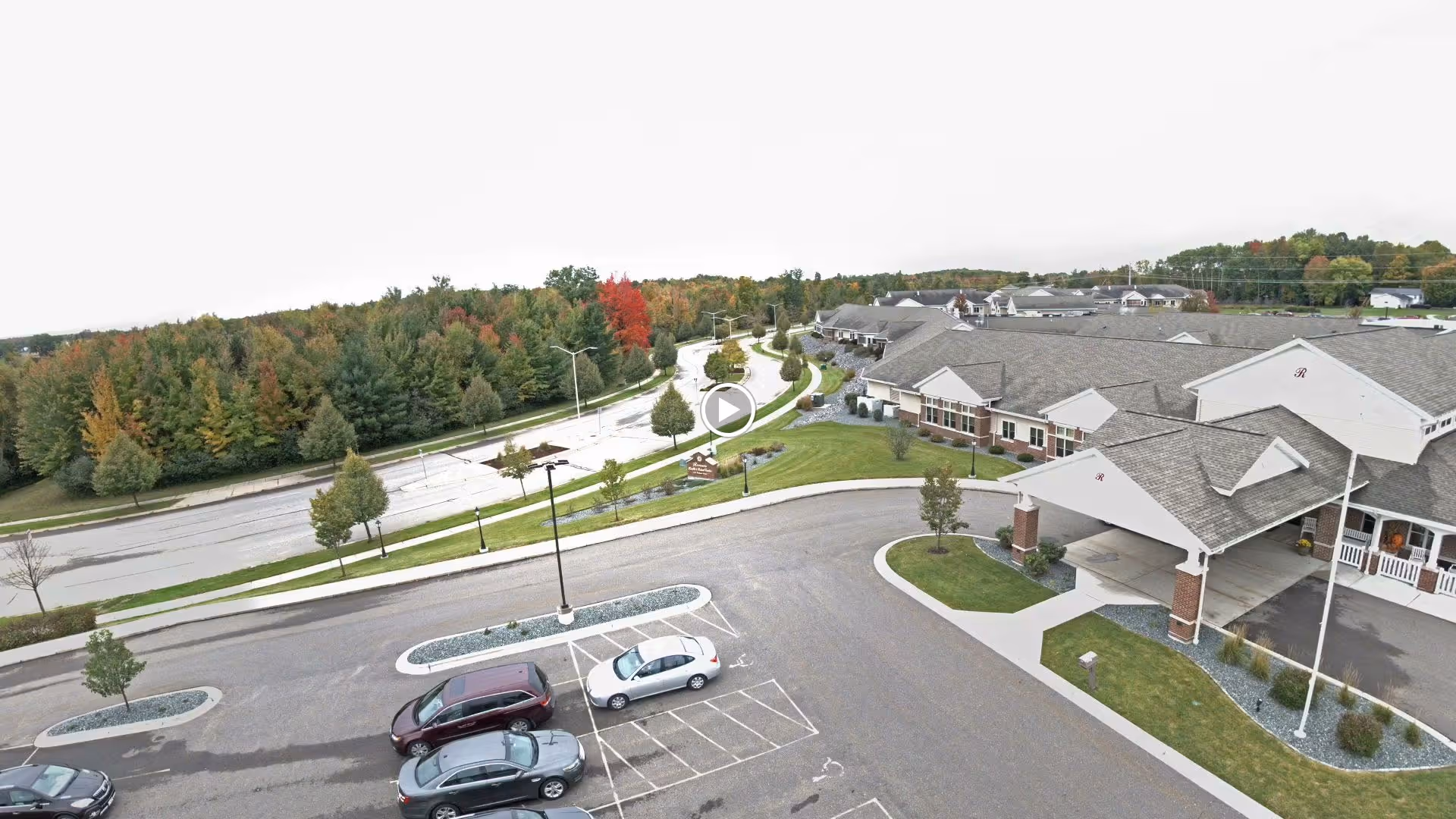 Aerial view of Rennes Health & Rehab Center - Peshtigo West showing the building with a covered entrance, parking lot with several cars, landscaped greenery, and a road lined with trees displaying autumn foliage.