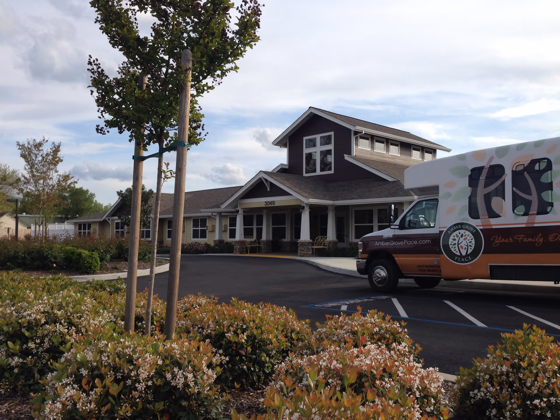 Exterior view of Amber Grove Place Memory Care facility showing a single-story building with a peaked roof, surrounded by landscaped bushes and young trees. A facility shuttle van with the Amber Grove Place logo and website is parked in front of the building under a partly cloudy sky.