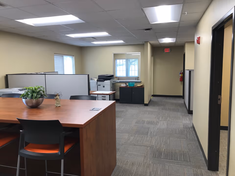 Office area with a wooden desk, potted plant, cubicles, and printers; hallway beyond.