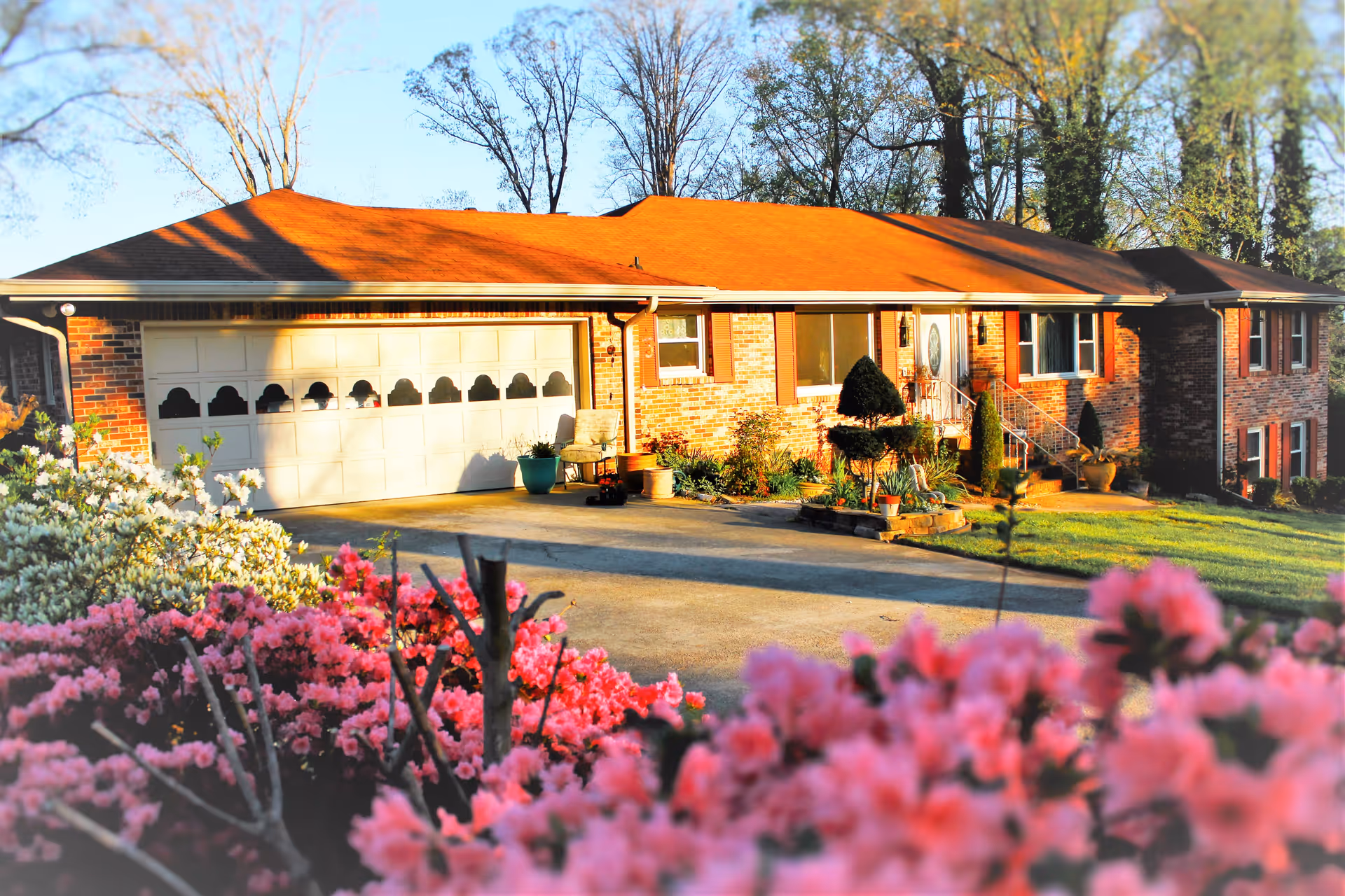Front exterior of a single-story brick house with a garage, driveway, and pink flowering shrubs in the foreground.