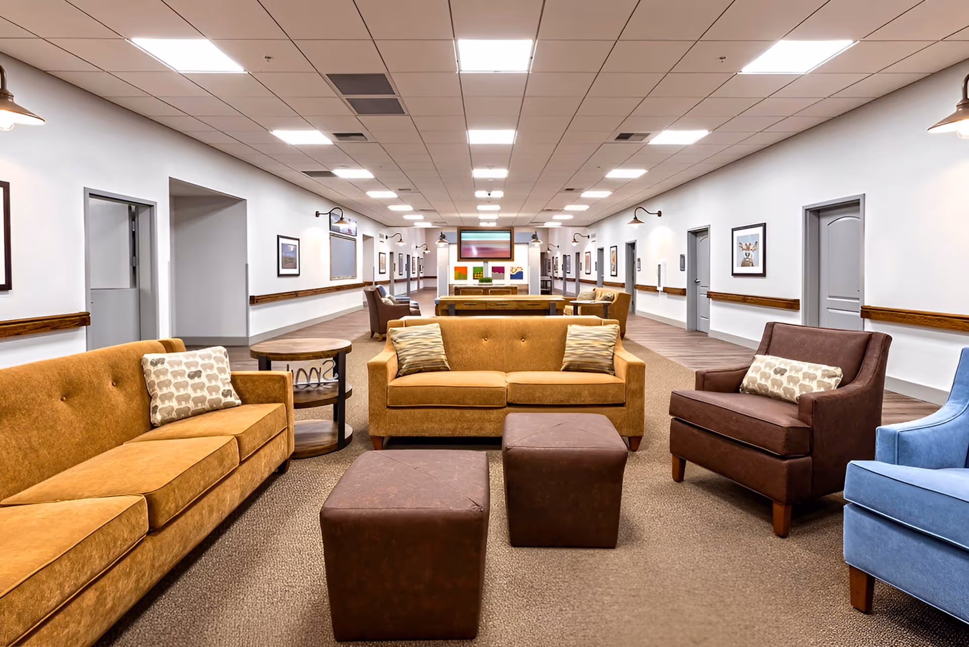 Spacious senior living lounge with tan sofas, chairs, and ottomans in a bright common area.