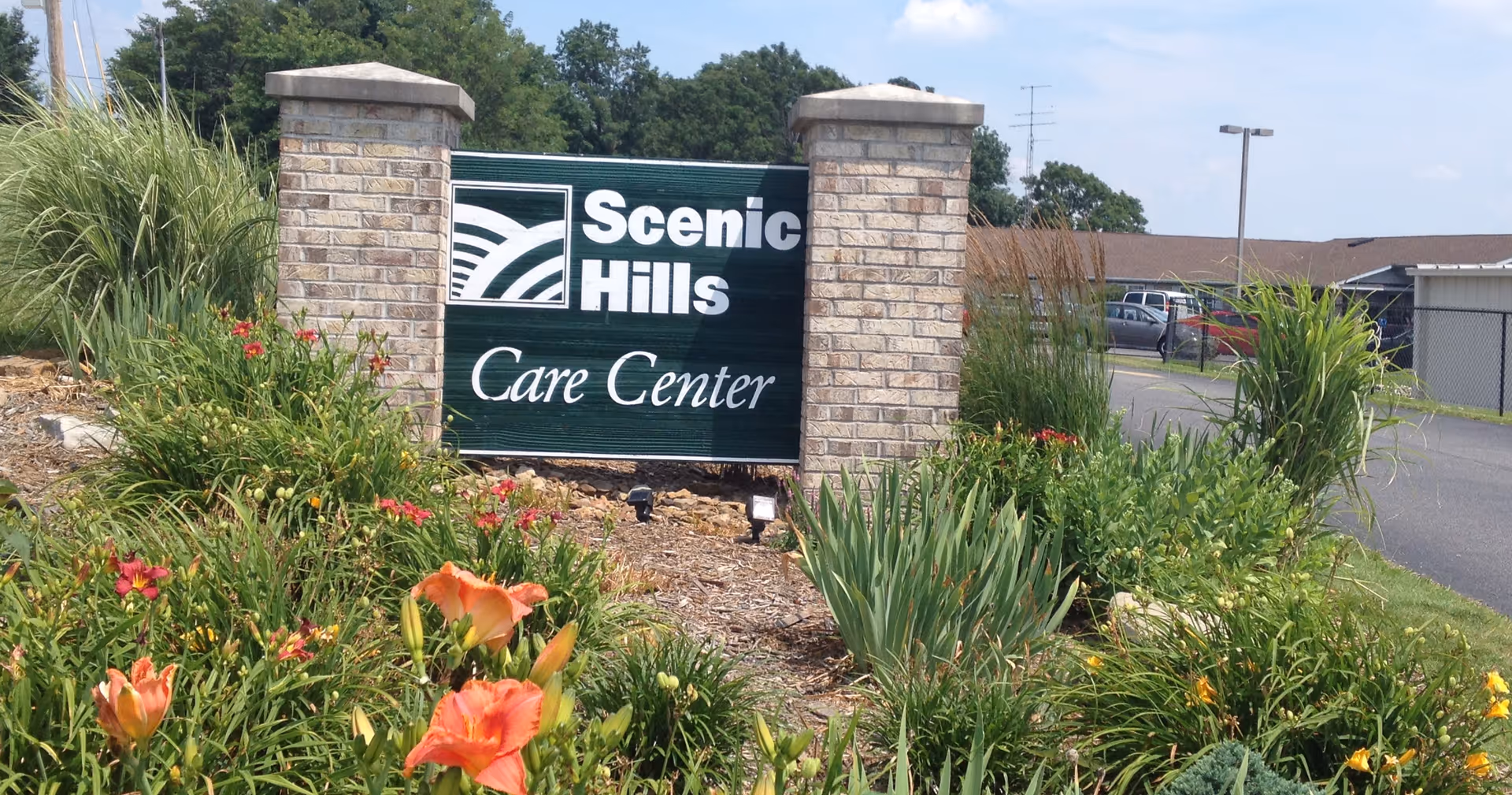 A landscaped entrance sign for Scenic Hills Care Center, featuring a green sign with white text and a logo, flanked by two brick pillars, surrounded by various green plants and orange flowers, with a parking lot and building visible in the background under a blue sky.