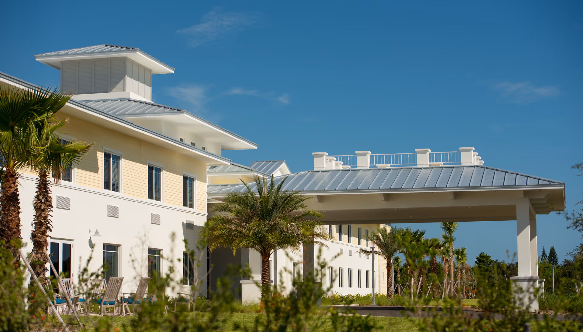 Front exterior of a low-rise senior living building with a covered porte-cochère, palm trees, and outdoor seating under a clear blue sky.