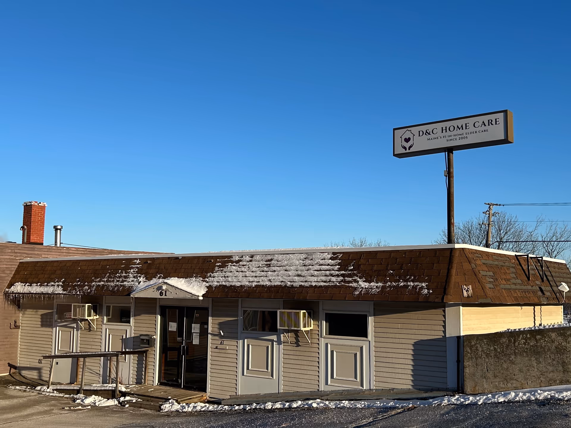 Exterior front view of D&C Home Care building with snow on roof under clear blue sky.