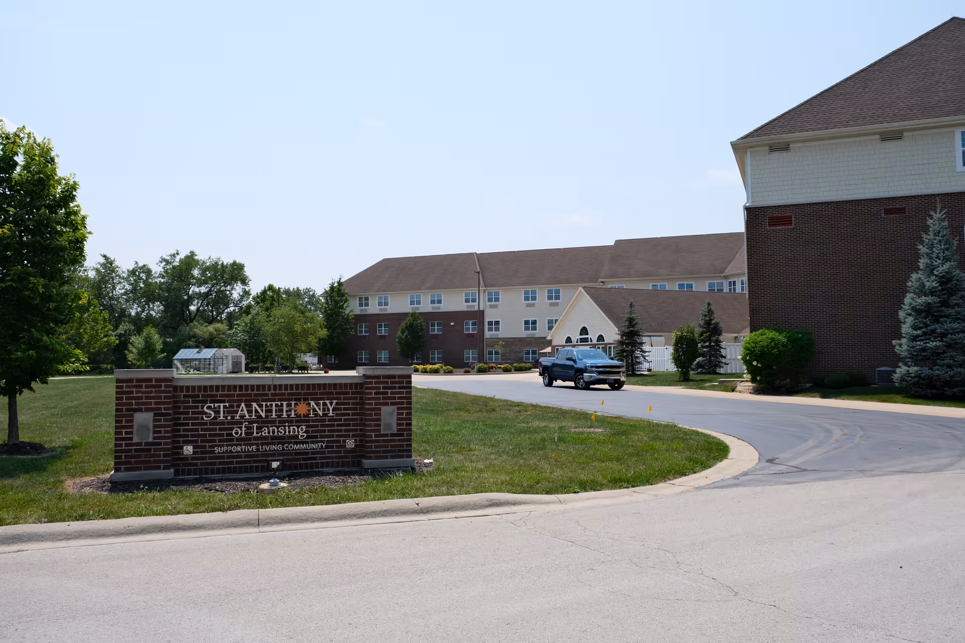 Exterior view of St. Anthony of Lansing supportive living community building with a brick sign in the foreground and a pickup truck on the driveway. The building is surrounded by trees and grass under a clear blue sky.