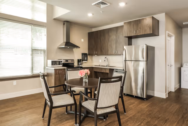 A bright modern kitchenette with a round dining table and four chairs, stainless steel appliances, and wood cabinets.
