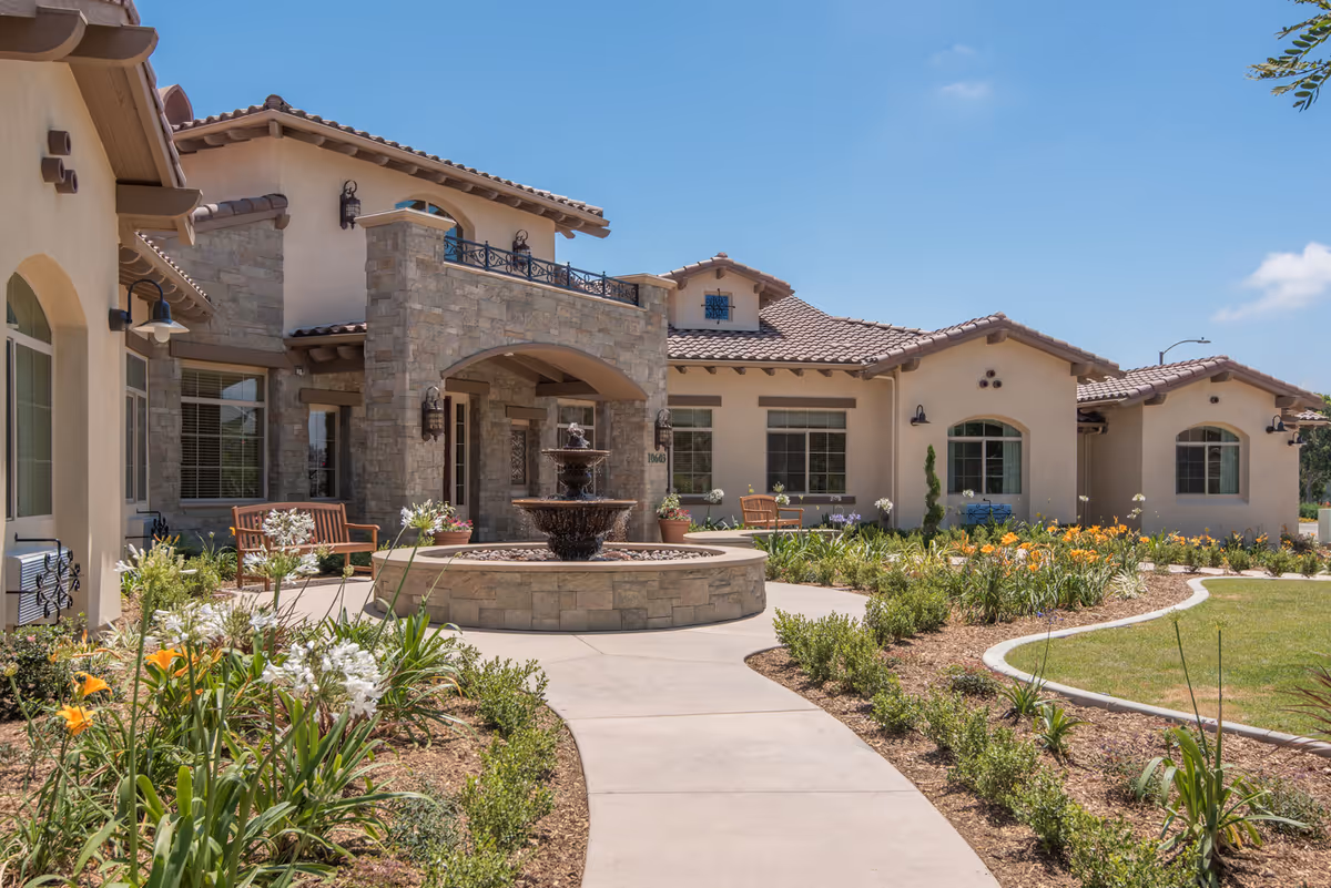 Exterior view of ActivCare 4S Ranch facility featuring a beige stucco building with stone accents, a tiled roof, and multiple windows. In front, there is a circular stone fountain surrounded by a landscaped garden with flowers and shrubs. A paved walkway leads to the entrance, and wooden benches are placed along the path under a clear blue sky.