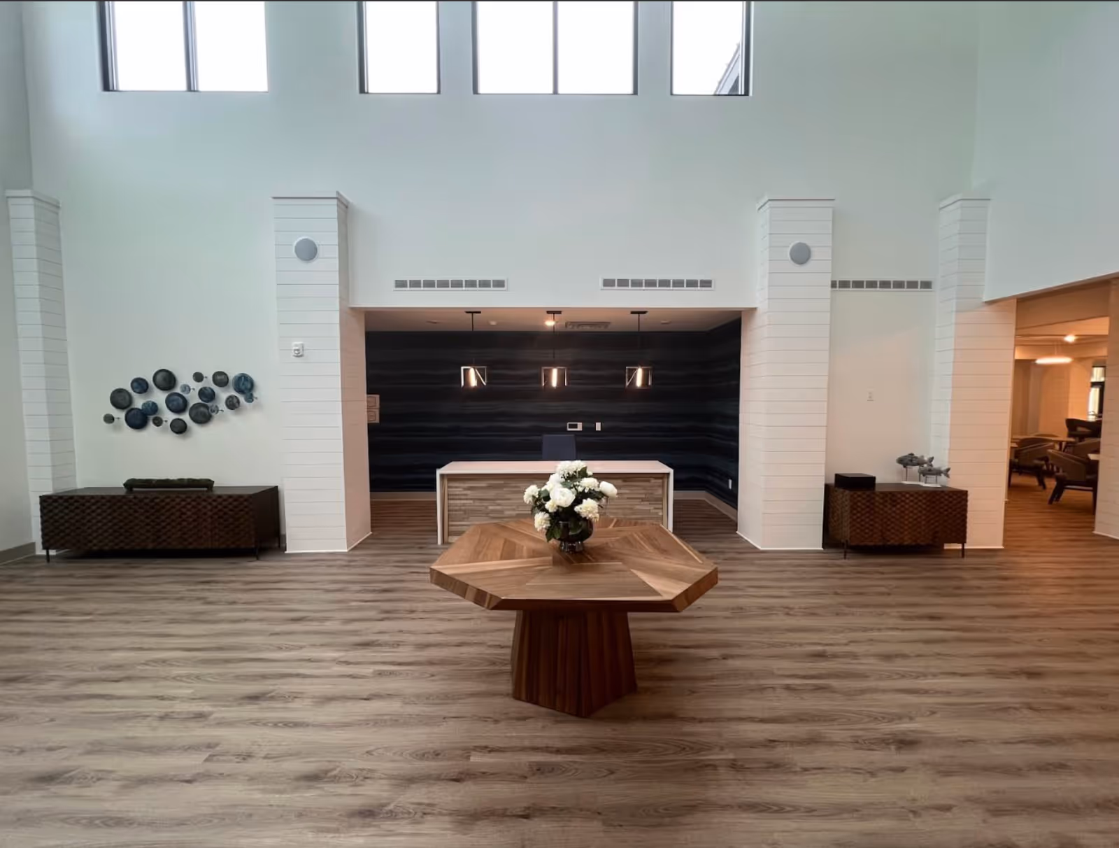 A spacious, modern lobby area with high ceilings and large windows near the ceiling. In the center is a wooden octagonal table with a vase of white flowers. Behind the table is a reception desk with three pendant lights hanging above it. On either side of the desk are wooden cabinets, one with decorative wall art above it and the other with small sculptures. The floor is wood-patterned, and there is an open doorway to the right leading to a seating area with chairs.