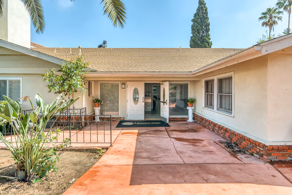 Front entrance of a single-story assisted living building with a covered porch, potted plants, and a welcome mat.