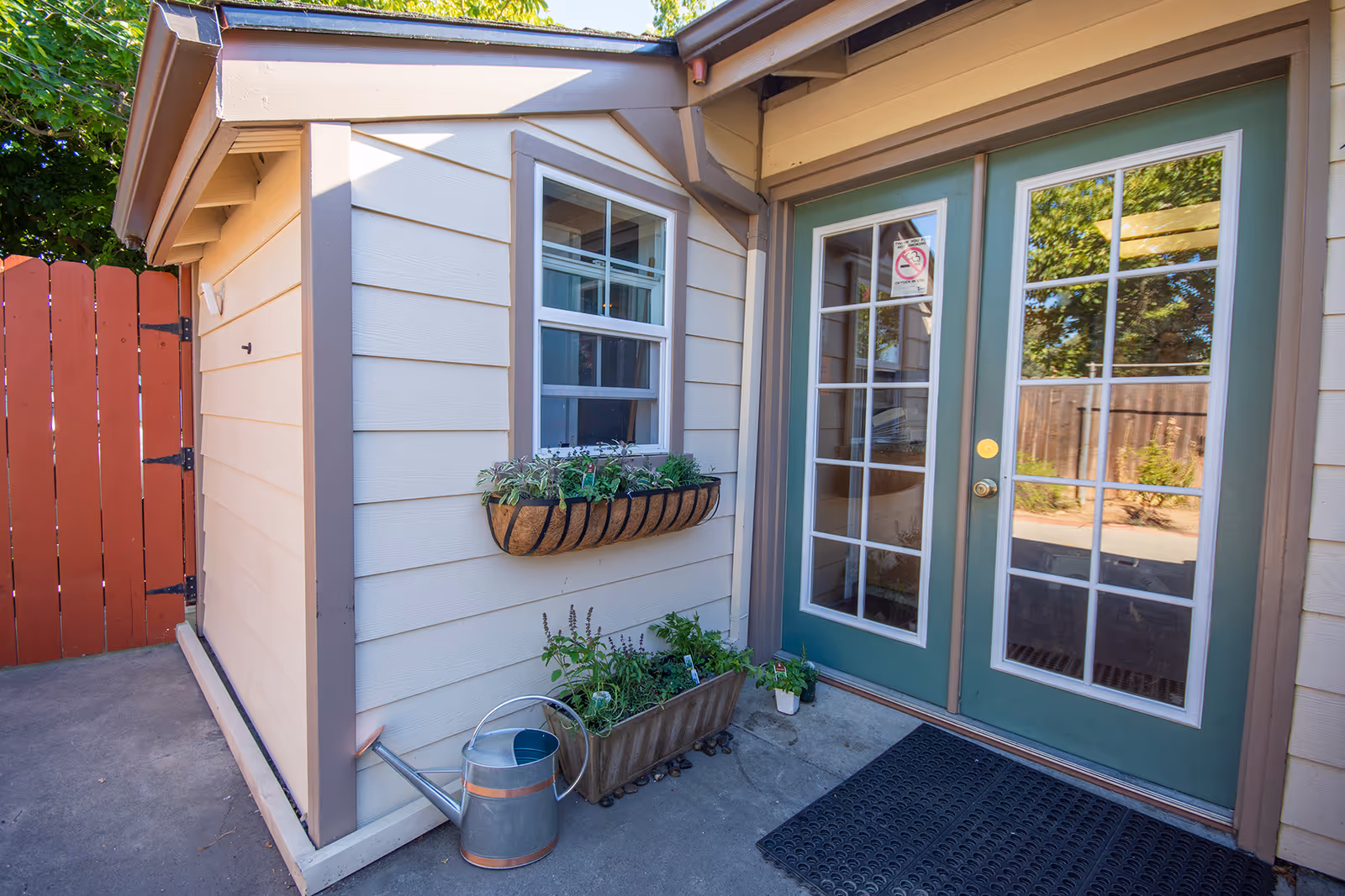 Exterior view of a beige building entrance with green French doors, window box, and plants.