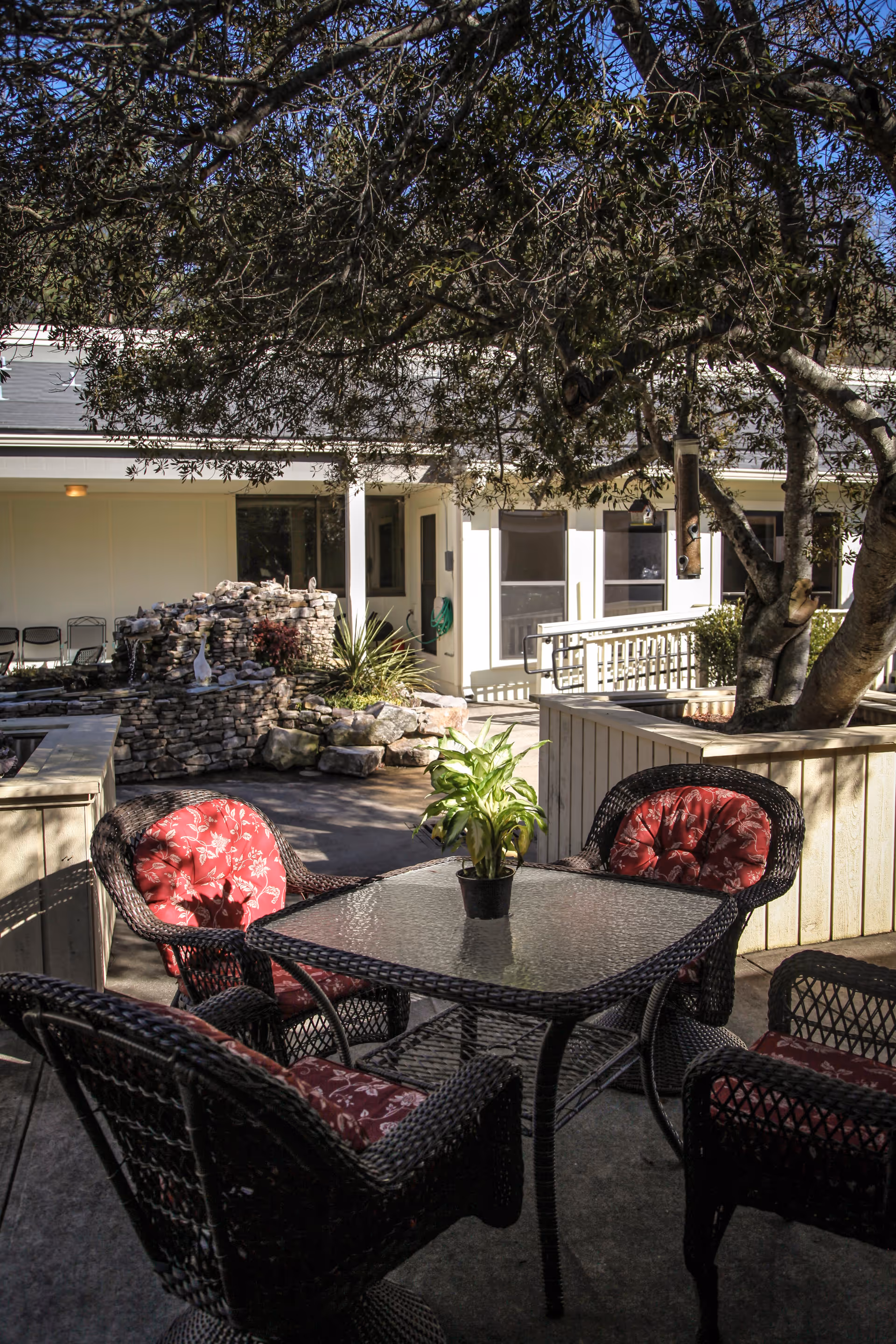 Outdoor patio area with a glass-top table surrounded by four wicker chairs with red floral cushions. A small potted plant is on the table. In the background, there is a stone water feature and a building with windows and a ramp. Trees provide shade over the seating area.