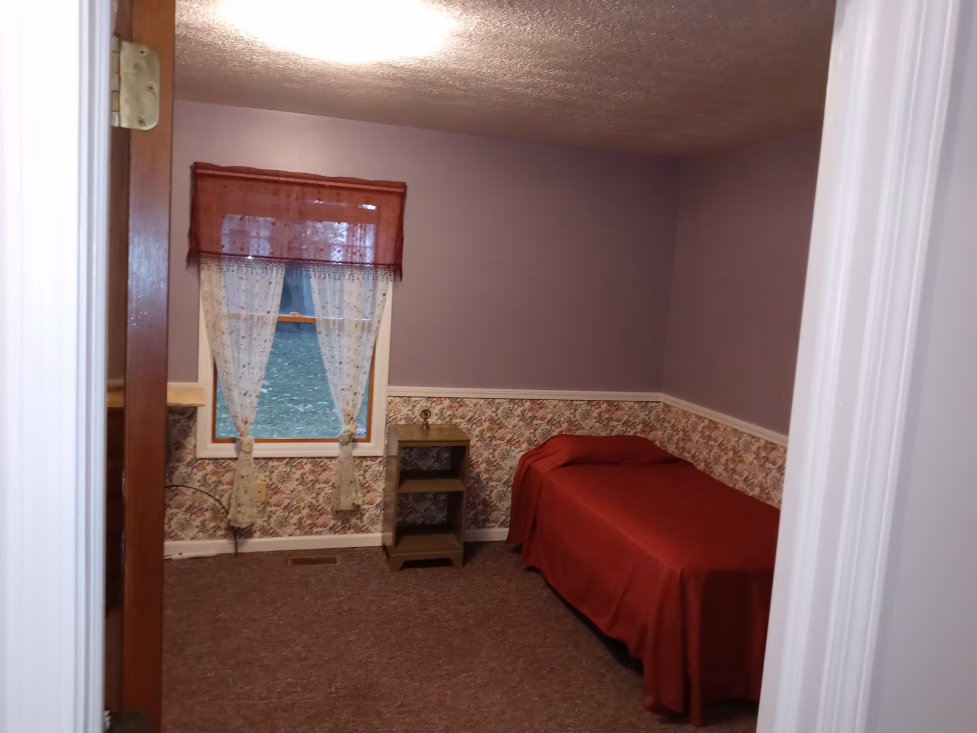 Small bedroom with purple walls, red bedspread, floral wallpaper, and window with curtains.