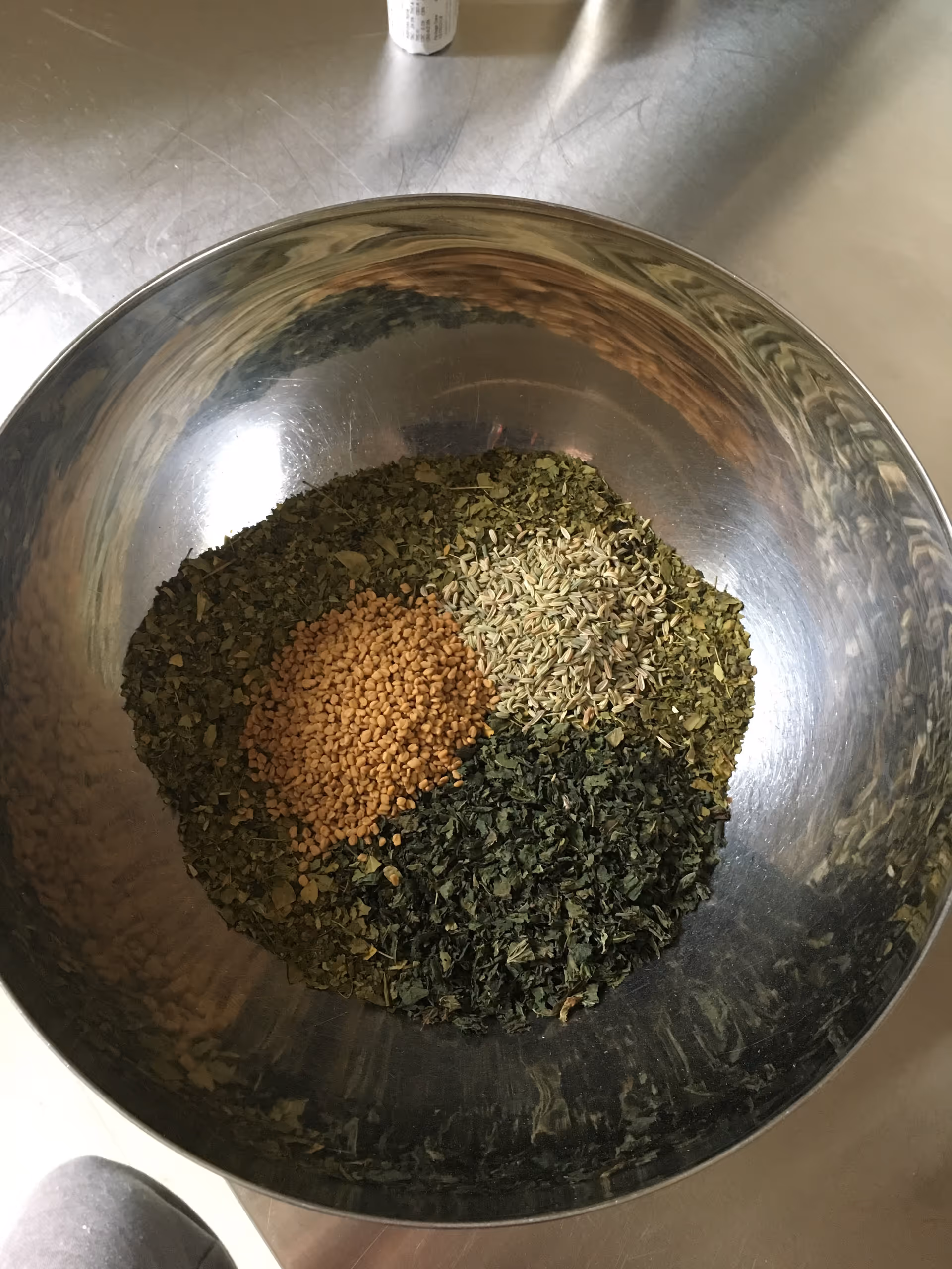 A stainless steel bowl holds assorted dried herbs and seeds on a stainless kitchen counter.