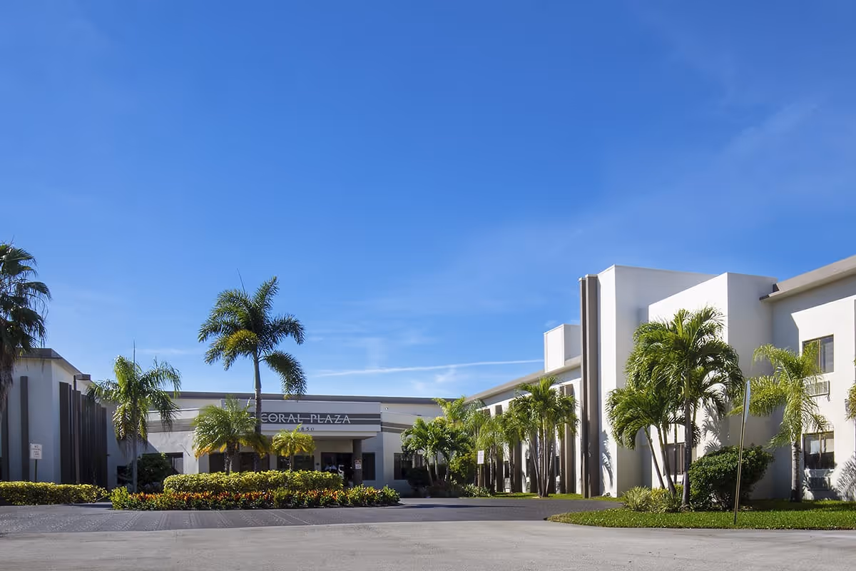 Exterior front view of Courtyard Plaza facility with white buildings, palm trees, and a clear blue sky.