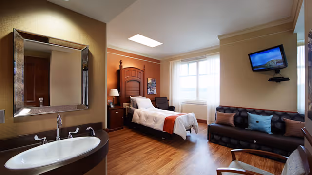 Hotel-style senior living suite with a bed, sofa, TV, and sink area.