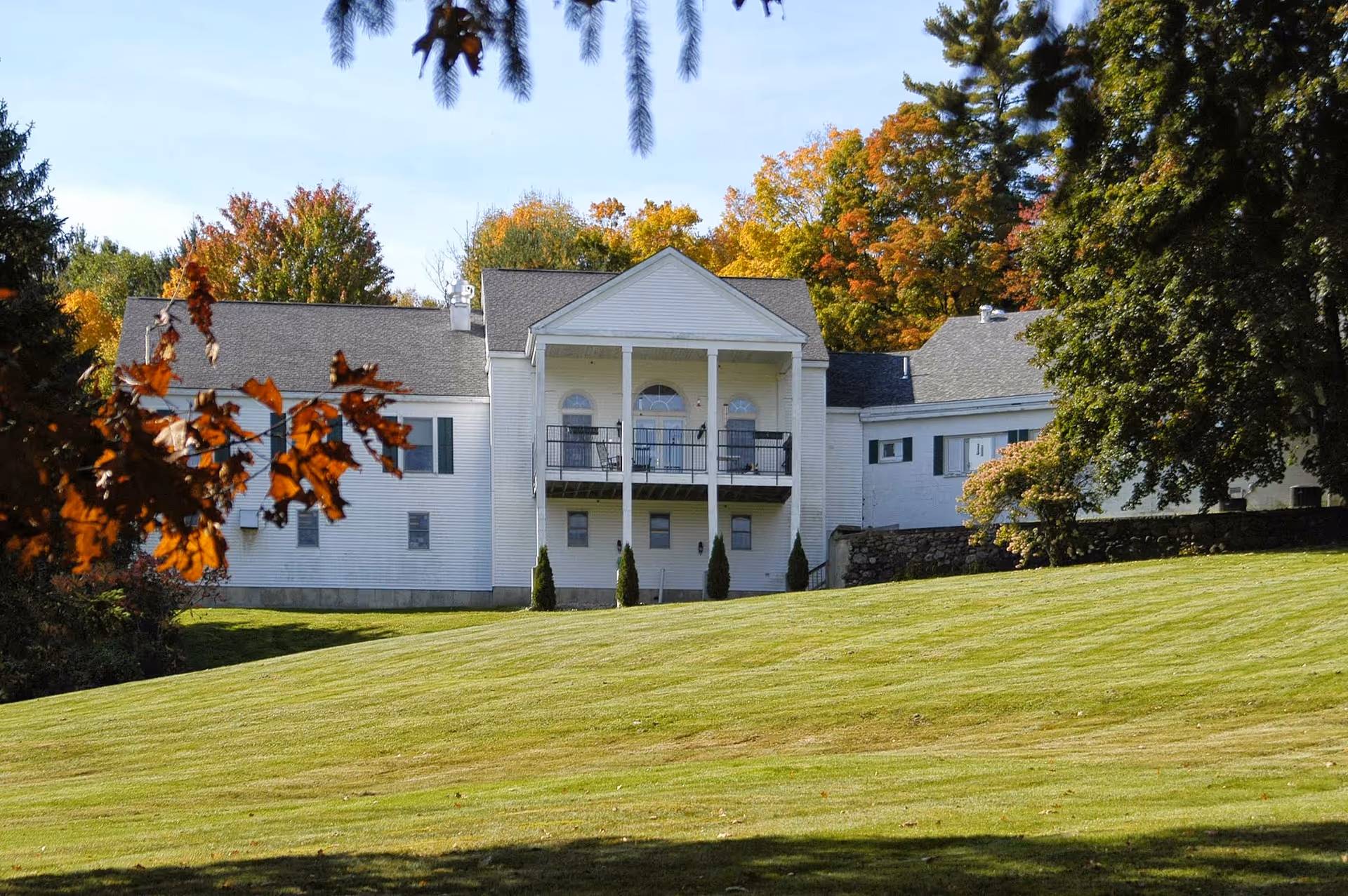 A large white building with a balcony and columns, surrounded by green grass and trees with autumn foliage under a clear blue sky.