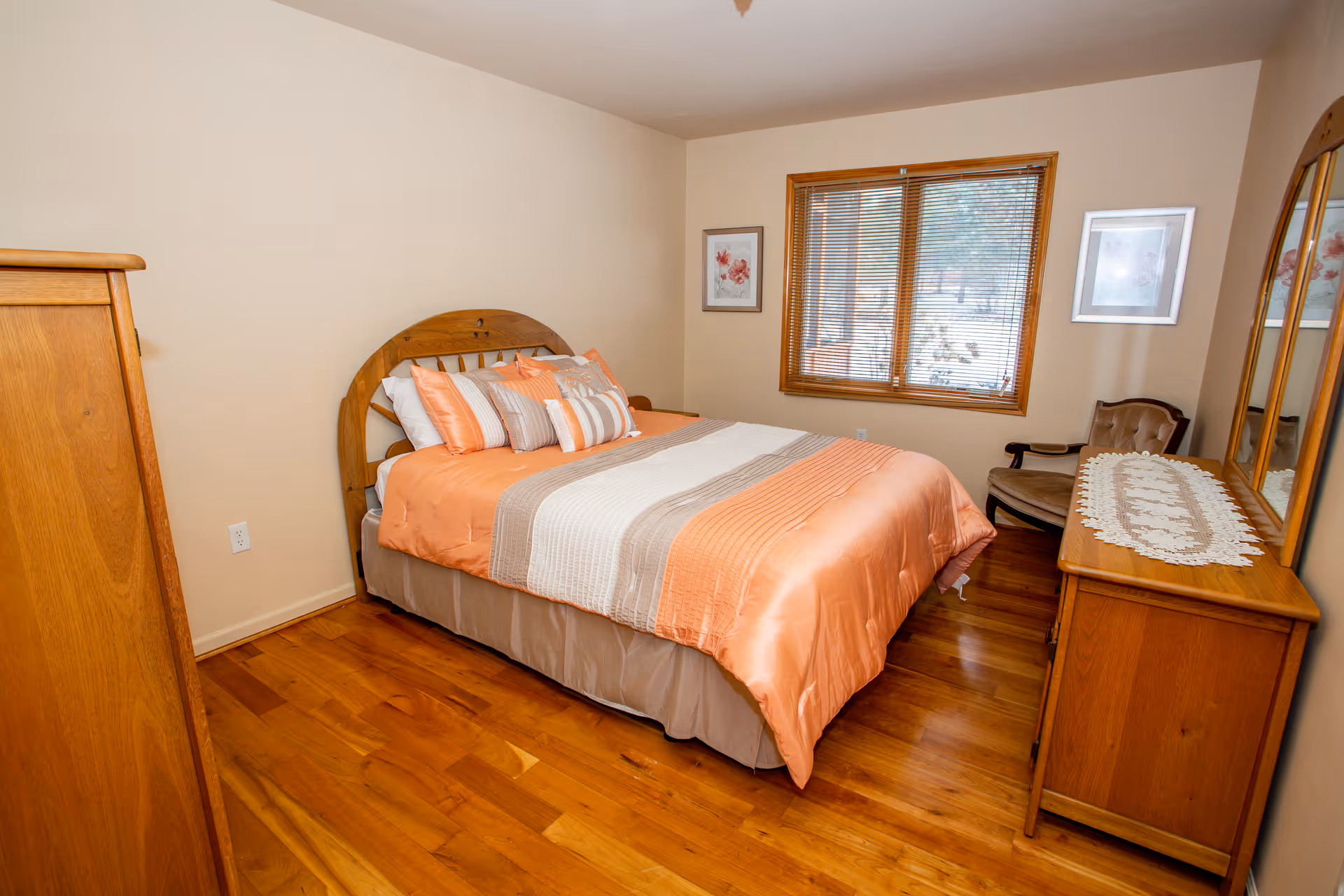 A cozy bedroom with a wooden bed frame and a neatly made bed featuring orange and beige bedding. The room has hardwood floors, a wooden dresser with a lace runner and mirror, a cushioned chair, and two framed floral pictures on the walls. A window with wooden blinds lets in natural light.