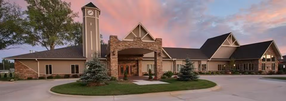 Exterior view of a senior living facility building with a clock tower, stone and beige walls, and a driveway with landscaped greenery under a colorful evening sky.